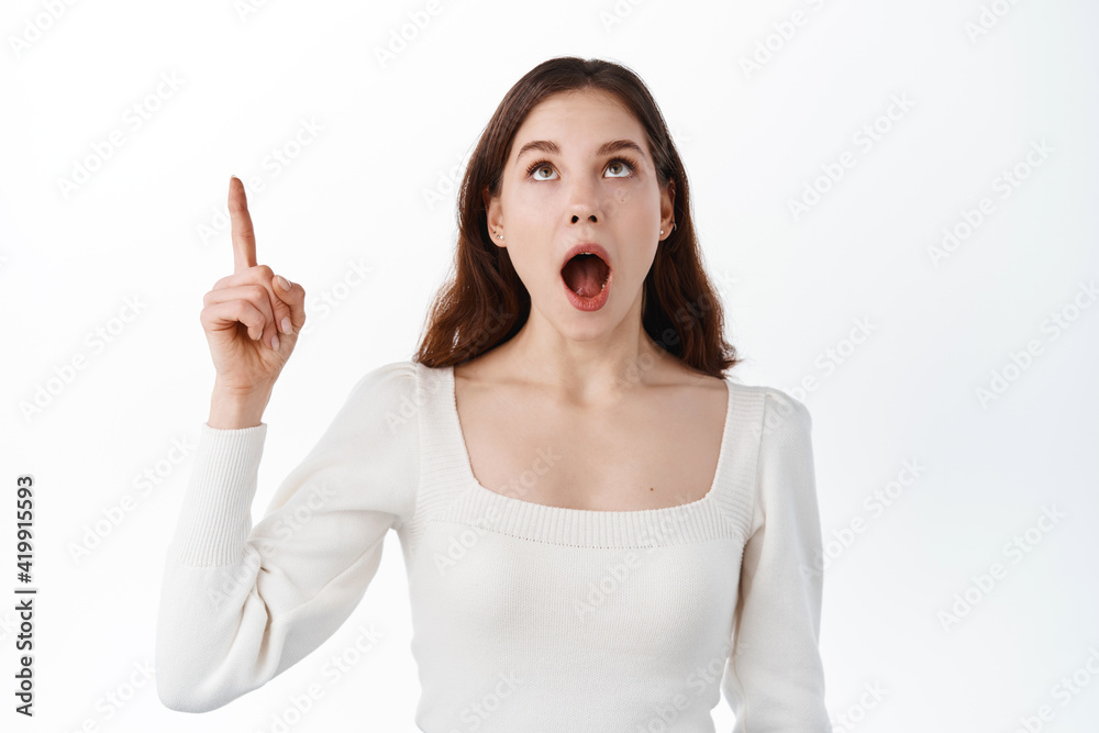 Surprised and impressed brunette girl drop jaw, look and pointing up at super cool promo offer, standing amazed with advertisement against white background Surprised and impressed brunette girl drop jaw, look and pointing up at super cool promo offer, standing amazed with advertisement against white background