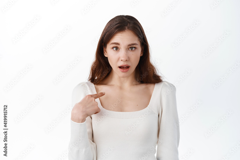 Confused girl pointing at herself with surprised face, being chosen or accused, stare puzzled at camera, standing against white studio background Confused girl pointing at herself with surprised face, being chosen or accused, stare puzzled at camera, standing against white studio background