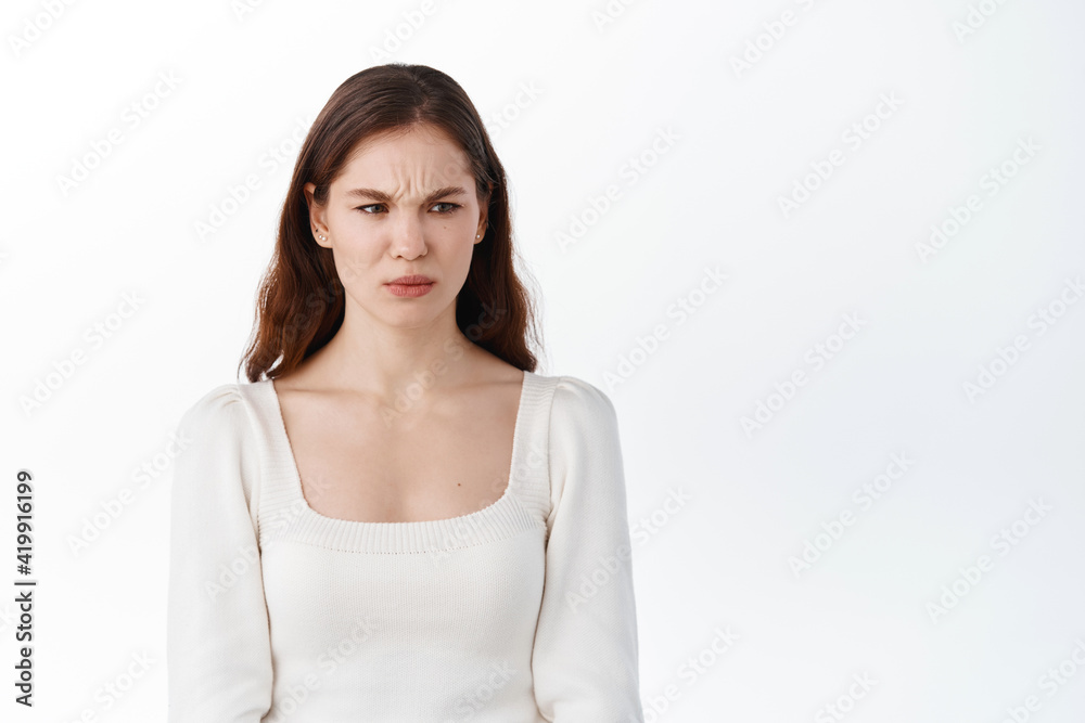 Frowning displeased woman looking aside at something awful or disgusting, express disgust and aversion, dislike bad product, standing against white background Frowning displeased woman looking aside at something awful or disgusting, express disgust and aversion, dislike bad product, standing against white background