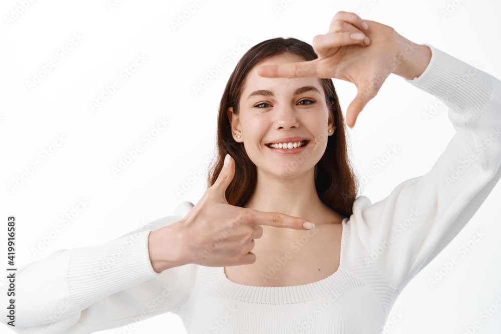 Portrait of beautiful young girl with white smile, smiling face, showing hand camera frames and looking through with pleased expression, standing over white background Portrait of beautiful young girl with white smile, smiling face, showing hand camera frames and looking through with pleased expression, standing over white background