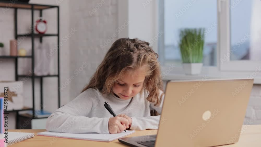 Cute primary school girl studying at home using laptop computer. Schoolgirl has online lesson, writing in exercise book, doing homework. Distance learning online. Children home education Cute primary school girl studying at home using laptop computer. Schoolgirl has online lesson, writing in exercise book, doing homework. Distance learning online. Children home education