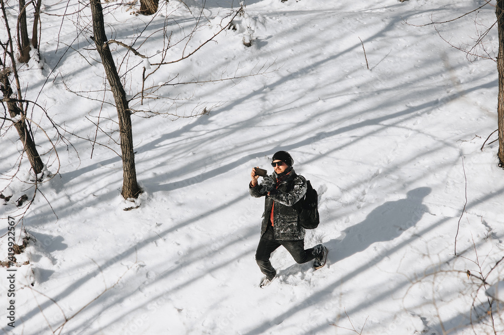 An adult man, a wanderer, a tourist in black warm clothes and a large backpack on his back travels through the snowy forest and takes pictures of the beautiful nature on a mobile phone, smartphone.