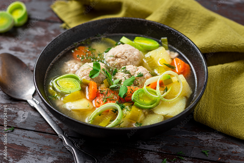 Rustic soup with vegetables and meatballs in a bowl on a dark wooden table. Soup with meatballs, leeks, potatoes, carrots and herbs. Tasty lunch or dinner rustic style.