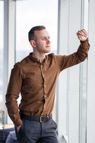 A young successful male businessman stands loosely near a large window and looks out over the city.
