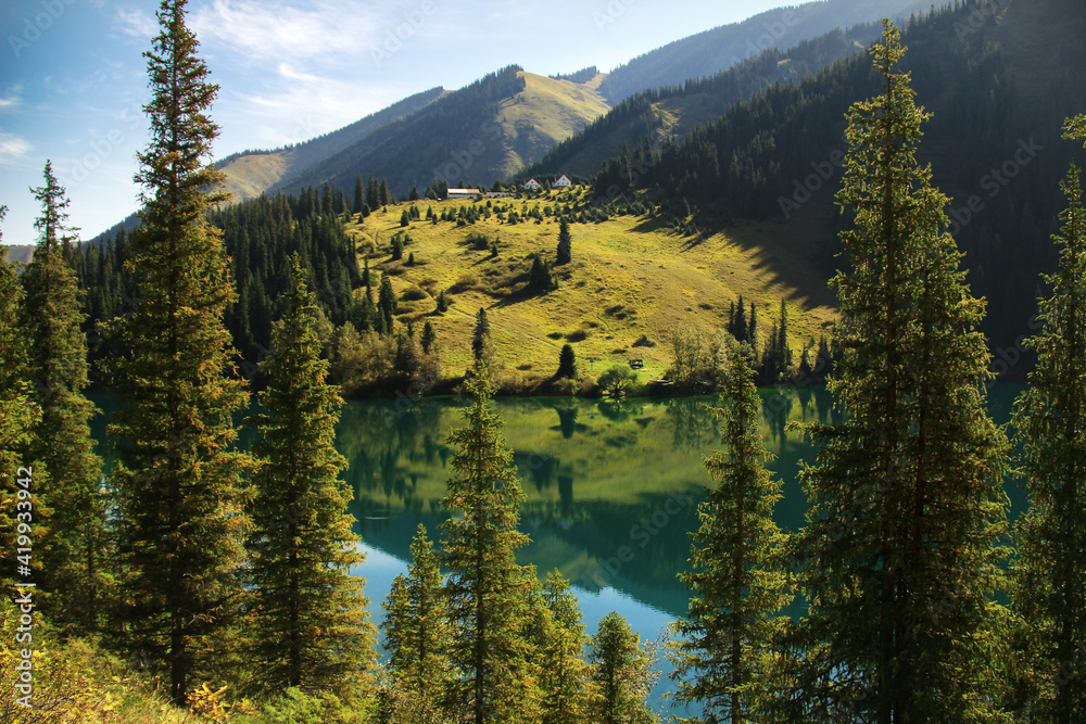 Alpine lake Kolsai, spruce and pine trees grow on the slopes, houses on the hills in the distance, mountains and trees are reflected in the water, summer, sunny, sky with clouds