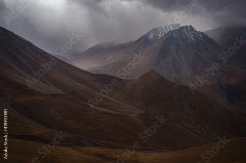 Autumn in the mountains, in a mountain valley, rain begins, the sky is in dark thick low clouds, the mountain slopes are covered with yellow grass, in the distance sunlight through the clouds