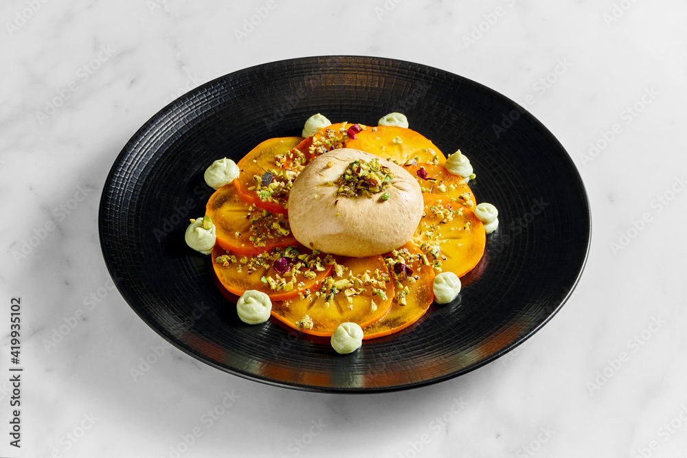 Buratta cheese with persimmon and pistachio in a black plate on a light marble background Buratta cheese with persimmon and pistachio in a black plate on a light marble background