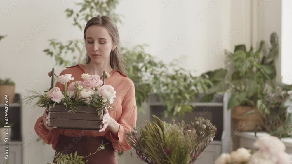 Medium POV of young Caucasian woman standing in plant shop, holding wooden basket with flowers and greens arrangement, looking and smiling on camera Medium POV of young Caucasian woman standing in plant shop, holding wooden basket with flowers and greens arrangement, looking and smiling on camera