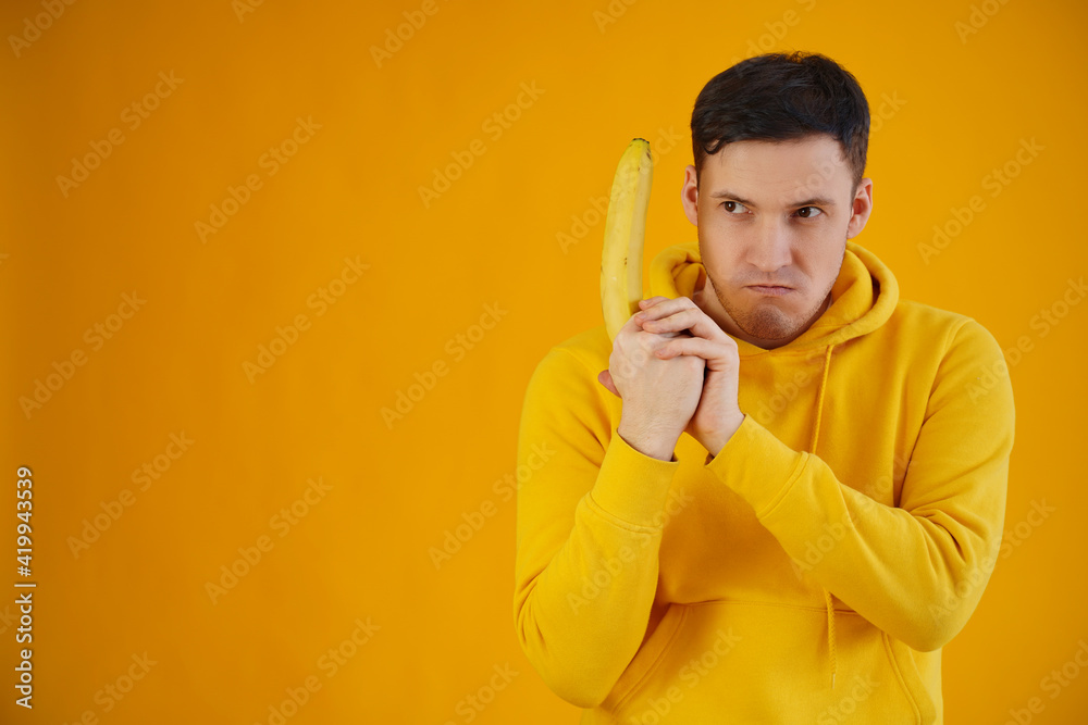 Portrait of young man with banana on yellow background. Close up of handsome guy in yellow hoodie plays with fruit, imagining it as weapon. Portrait of young man with banana on yellow background. Close up of handsome guy in yellow hoodie plays with fruit, imagining it as weapon.