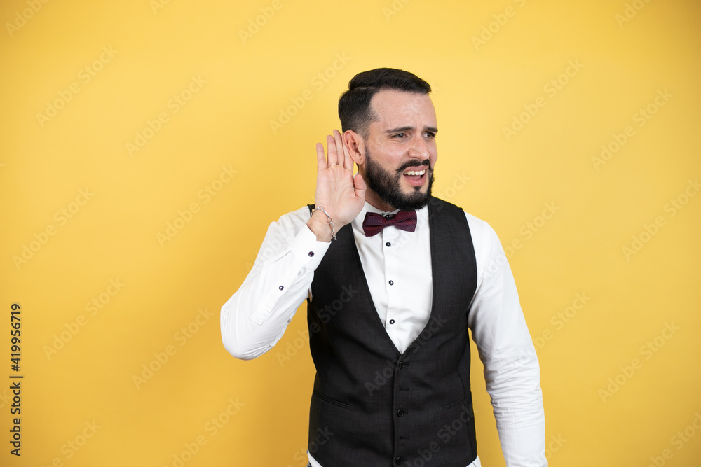 Young man with beard wearing bow tie and vest surprised with hand over ear listening an hearing to rumor or gossip Young man with beard wearing bow tie and vest surprised with hand over ear listening an hearing to rumor or gossip