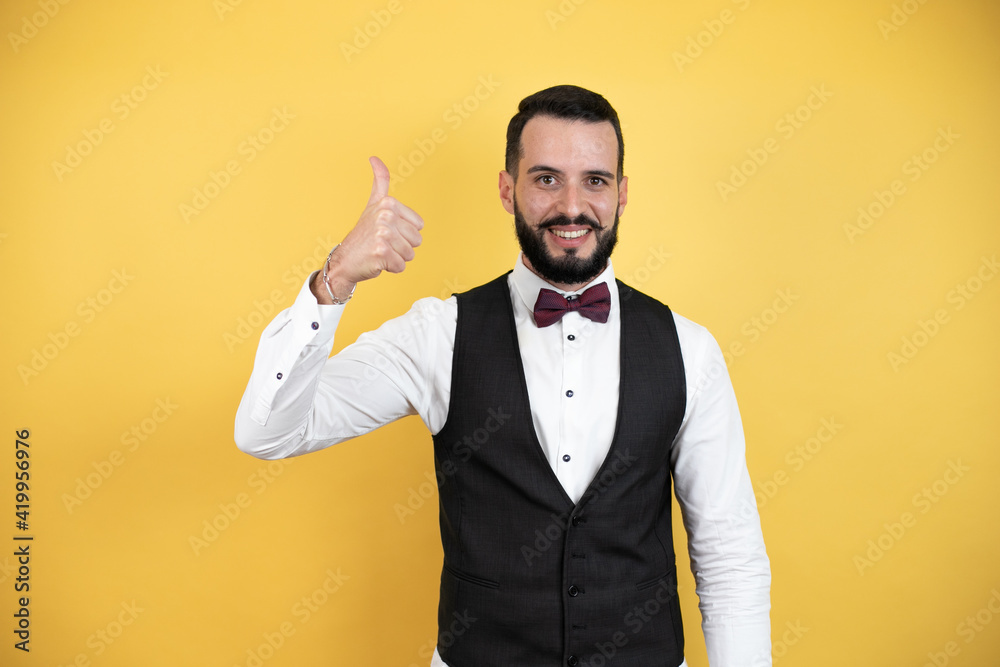 Young man with beard wearing bow tie and vest doing positive gesture with hand, thumbs up smiling and happy. cheerful expression Young man with beard wearing bow tie and vest doing positive gesture with hand, thumbs up smiling and happy. cheerful expression