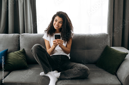 Modern stylish girl blogging at social media, using smartphone. Pretty african american woman chatting with friends or family and smiling while sitting at the couch in living room, browsing internet