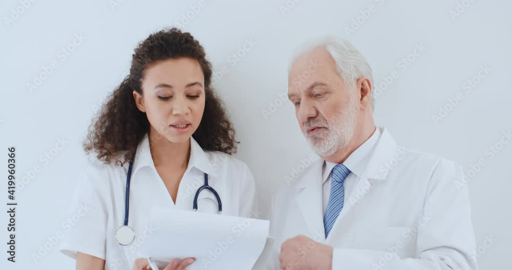 Portrait two medical workers aged caucasian male and young curly hair multiethnic female, doctors discussing medical case, talking about health report while standing by white wall in hospital Portrait two medical workers aged caucasian male and young curly hair multiethnic female, doctors discussing medical case, talking about health report while standing by white wall in hospital