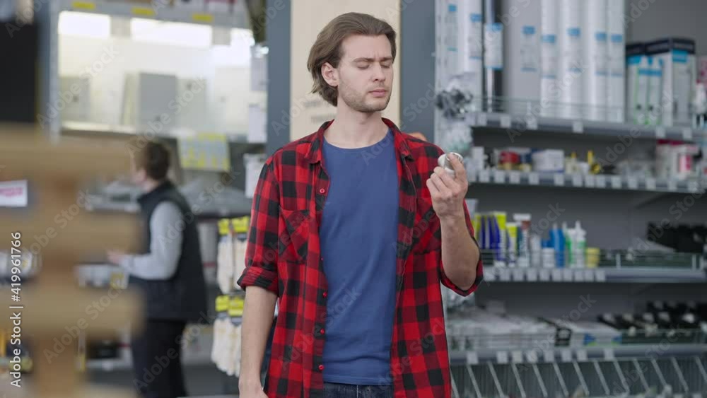 Thoughtful young man choosing appliances in hardware store. Blurred employee working at background between rows. Middle shot of confident concentrated Caucasian buyer making decision.