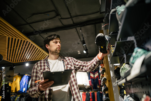 Business owner in sports store works with showcase trekking and mountaineering boots for sale, holding clipboard in his hands, checks availability of goods and collects to process online orders