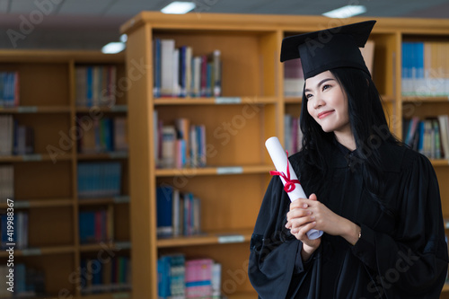 A young happy Asian woman university graduates in graduation gown and cap wears a face mask holds a degree certificate to celebrate her education achievement on the commencement day. Stock photo