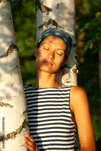 A girl in a vest and a bandana with closed eyes near a birch tree at sunset.