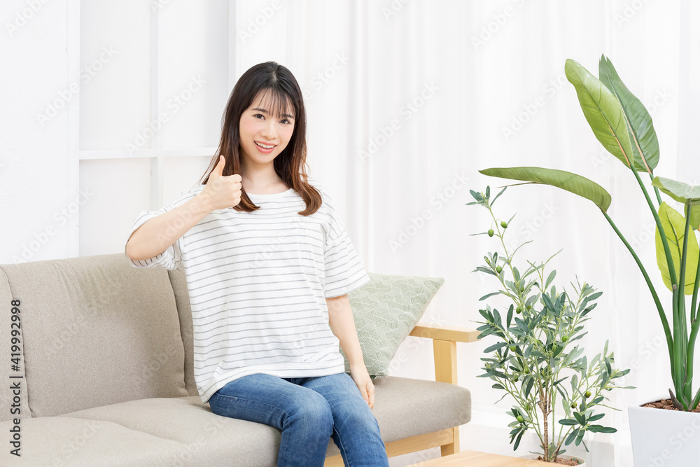 部屋のソファーに座って、いいねのジェスチャーをする若い女性 部屋のソファーに座って、いいねのジェスチャーをする若い女性