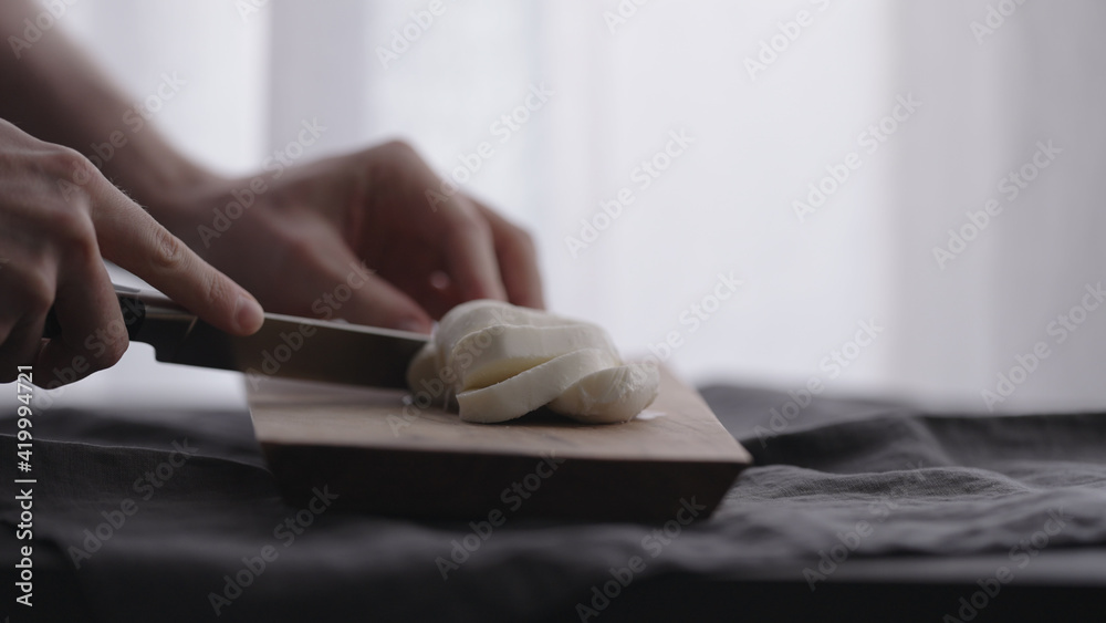 man slicing mozzarella ball on olive wood board with window on background