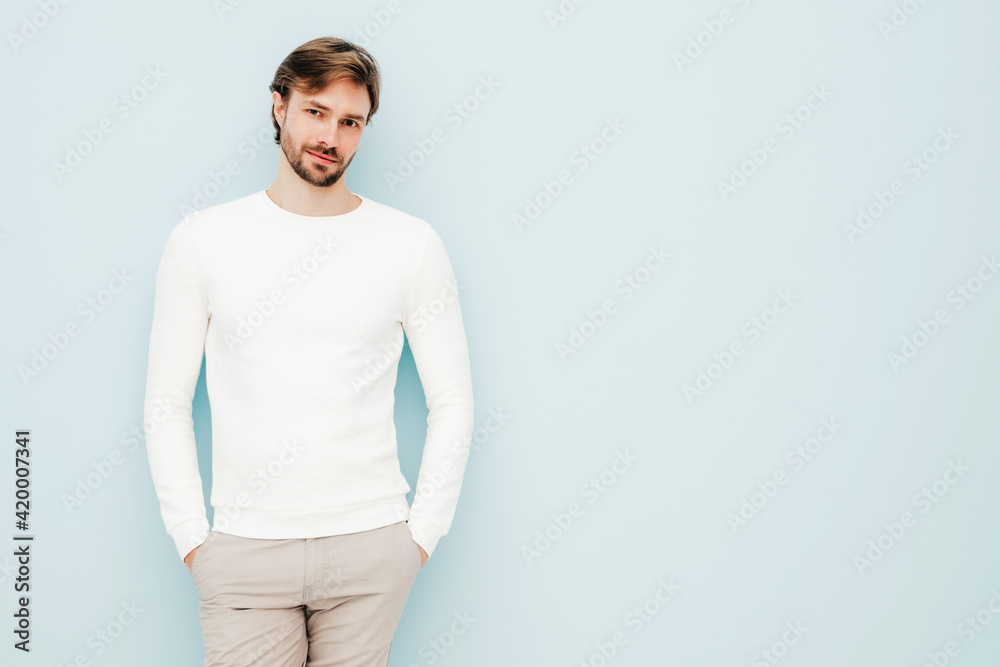 Portrait of handsome smiling hipster lumbersexual businessman model wearing casual white sweater and trousers. Fashion stylish man posing against light blue wall in studio Portrait of handsome smiling hipster lumbersexual businessman model wearing casual white sweater and trousers. Fashion stylish man posing against light blue wall in studio