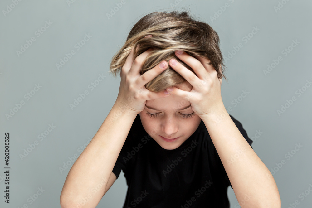 The sad boy sits at the table and has his head in his hands. Loneliness and stress. Grey background. Close-up. The sad boy sits at the table and has his head in his hands. Loneliness and stress. Grey background. Close-up.