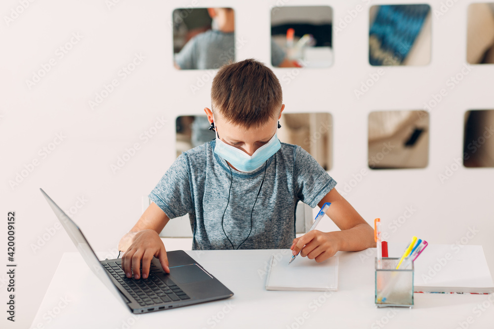 Young boy in headset sitting at table with laptop and medical face mask and preparing to school. Online education concept. Young boy in headset sitting at table with laptop and medical face mask and preparing to school. Online education concept.