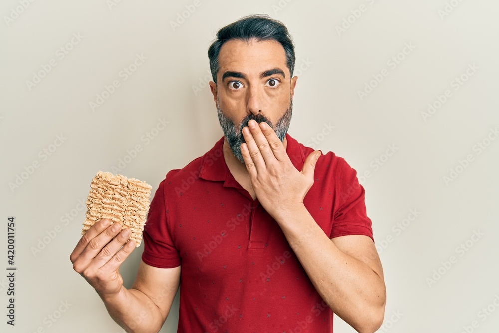 Middle age man with beard and grey hair holding uncooked noodles covering mouth with hand, shocked and afraid for mistake. surprised expression Middle age man with beard and grey hair holding uncooked noodles covering mouth with hand, shocked and afraid for mistake. surprised expression