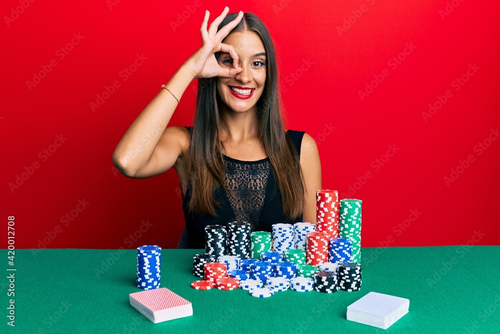 Young hispanic woman sitting on the table playing poker smiling happy doing ok sign with hand on eye looking through fingers Young hispanic woman sitting on the table playing poker smiling happy doing ok sign with hand on eye looking through fingers