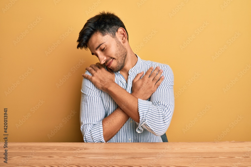 Young handsome man wearing casual clothes sitting on the table hugging oneself happy and positive, smiling confident. self love and self care Young handsome man wearing casual clothes sitting on the table hugging oneself happy and positive, smiling confident. self love and self care