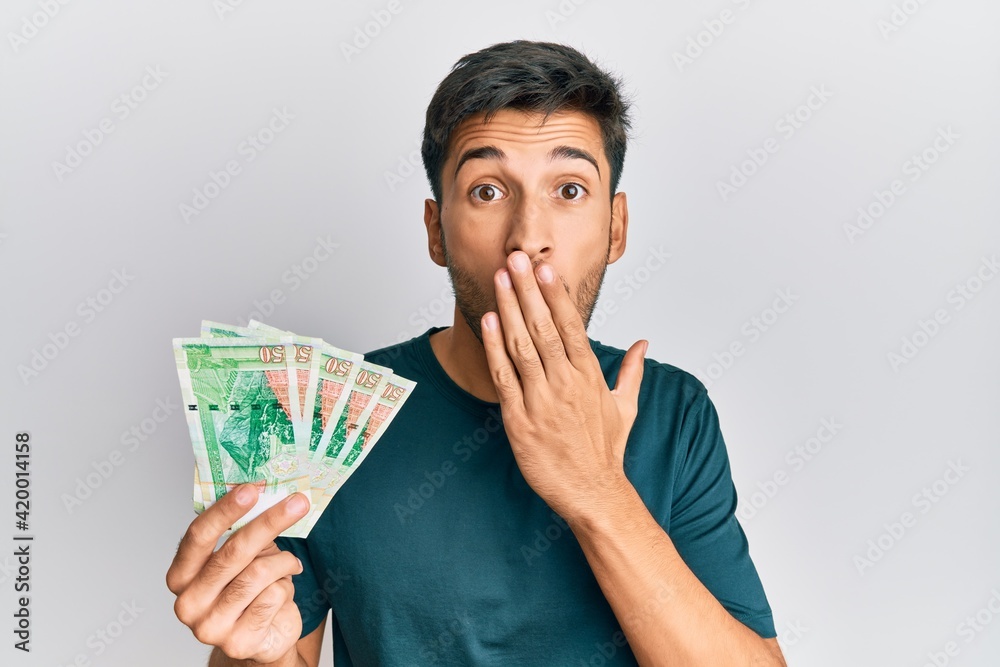 Young handsome man holding 50 hong kong dollars banknotes covering mouth with hand, shocked and afraid for mistake. surprised expression Young handsome man holding 50 hong kong dollars banknotes covering mouth with hand, shocked and afraid for mistake. surprised expression