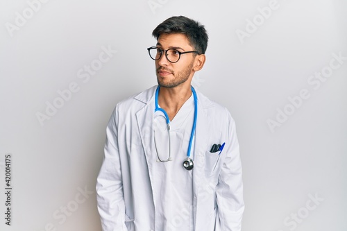 Young handsome man wearing doctor uniform and stethoscope smiling looking to the side and staring away thinking.