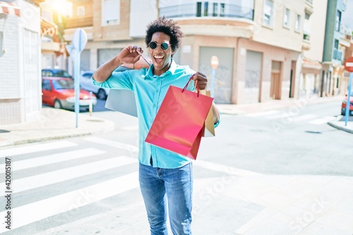 Young handsome african american man wearing sunglasses smiling happy. Standing with smile on face holding shopping bags at town street.