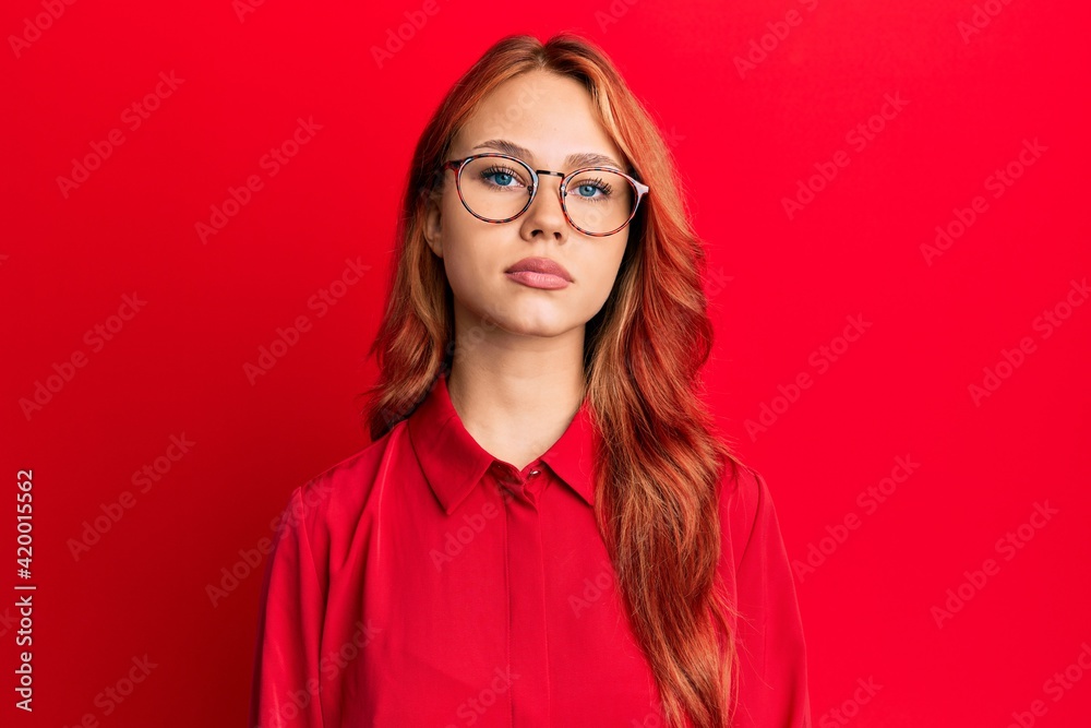 Young beautiful redhead woman wearing casual clothes and glasses over red background relaxed with serious expression on face. simple and natural looking at the camera.