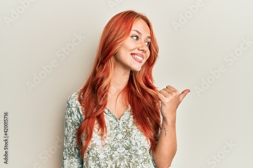 Young beautiful redhead woman wearing casual clothes smiling with happy face looking and pointing to the side with thumb up.