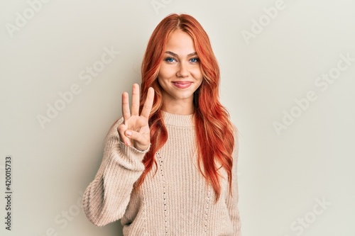 Young beautiful redhead woman wearing casual winter sweater showing and pointing up with fingers number three while smiling confident and happy.