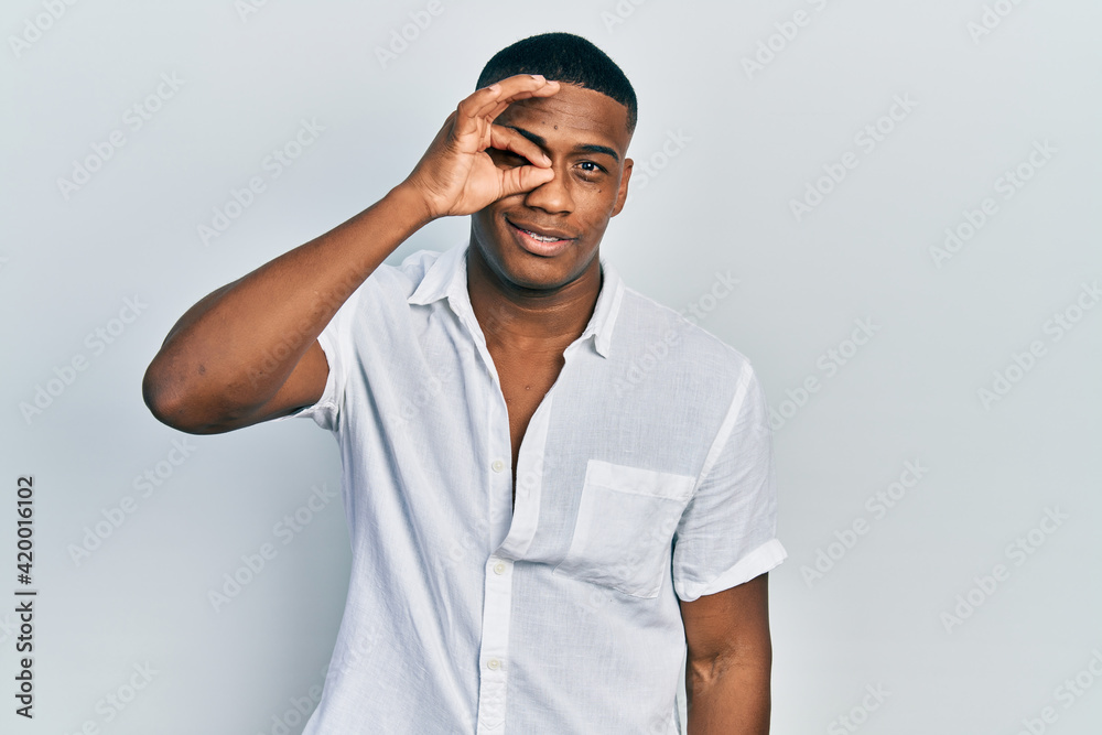 Young black man wearing casual white shirt doing ok gesture with hand smiling, eye looking through fingers with happy face. Young black man wearing casual white shirt doing ok gesture with hand smiling, eye looking through fingers with happy face.
