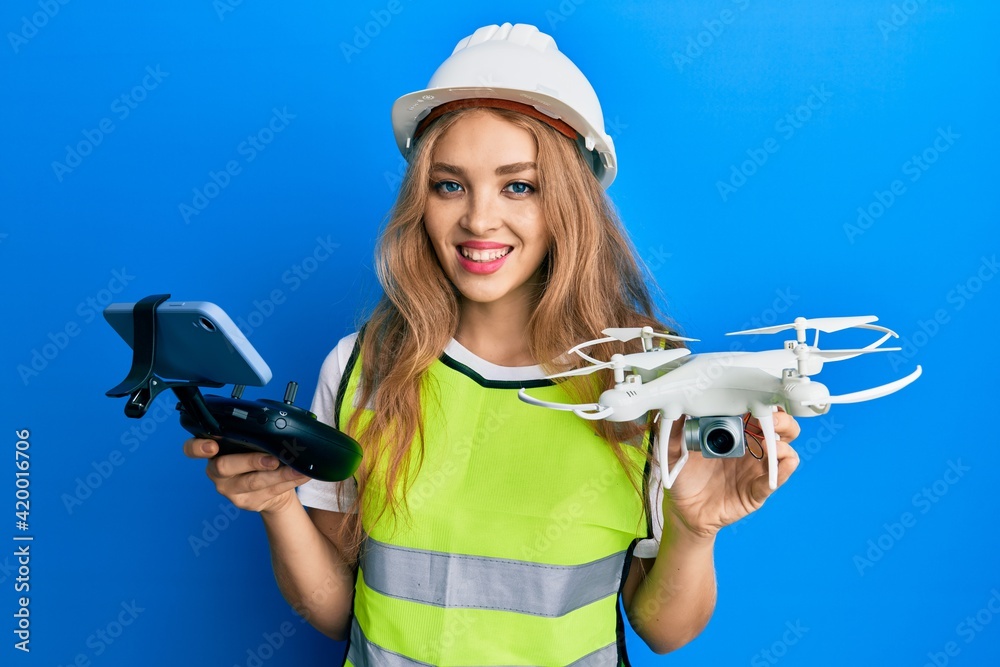 Beautiful blonde caucasian woman wearing safety hard using drone smiling with a happy and cool smile on face. showing teeth. Beautiful blonde caucasian woman wearing safety hard using drone smiling with a happy and cool smile on face. showing teeth.