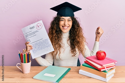 Young hispanic girl wearing graduated hat holding passed test screaming proud, celebrating victory and success very excited with raised arm