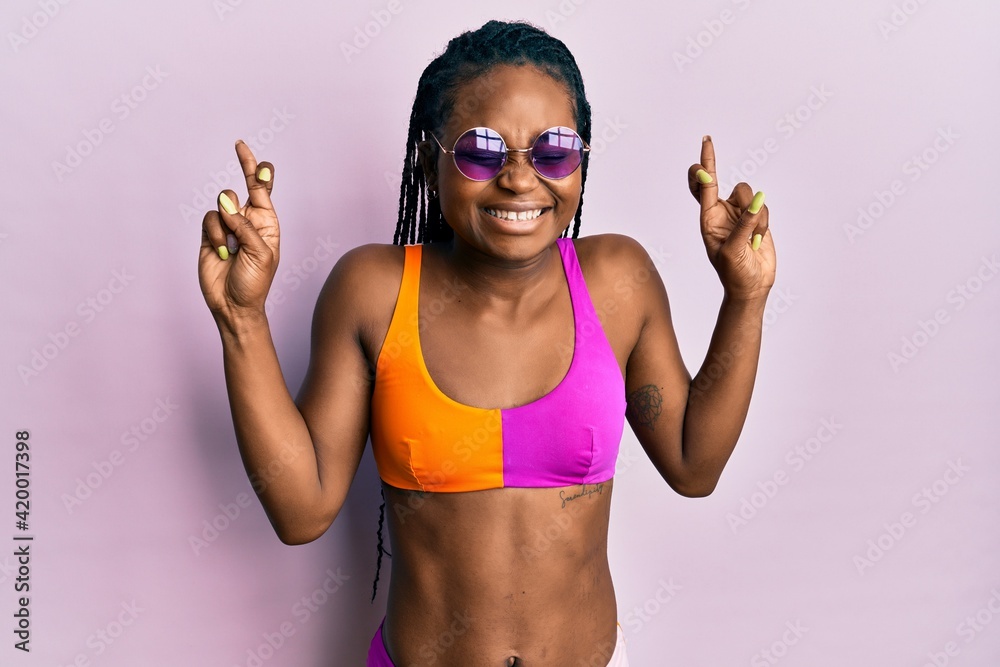 Young african american woman wearing bikini and sunglasses gesturing finger crossed smiling with hope and eyes closed. luck and superstitious concept.