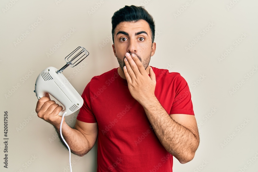 Young hispanic man holding food processor mixer machine covering mouth with hand, shocked and afraid for mistake. surprised expression Young hispanic man holding food processor mixer machine covering mouth with hand, shocked and afraid for mistake. surprised expression
