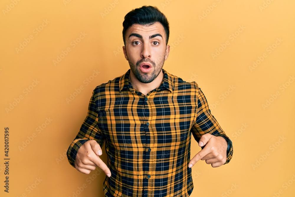 Young hispanic man wearing casual clothes pointing down with fingers showing advertisement, surprised face and open mouth