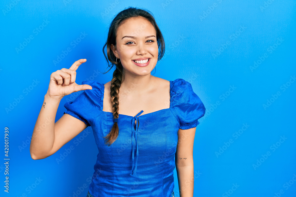 Young hispanic girl wearing casual clothes smiling and confident gesturing with hand doing small size sign with fingers looking and the camera. measure concept. Young hispanic girl wearing casual clothes smiling and confident gesturing with hand doing small size sign with fingers looking and the camera. measure concept.
