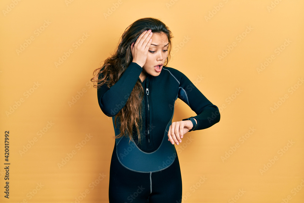 Young hispanic girl wearing diver neoprene uniform looking at the watch time worried, afraid of getting late