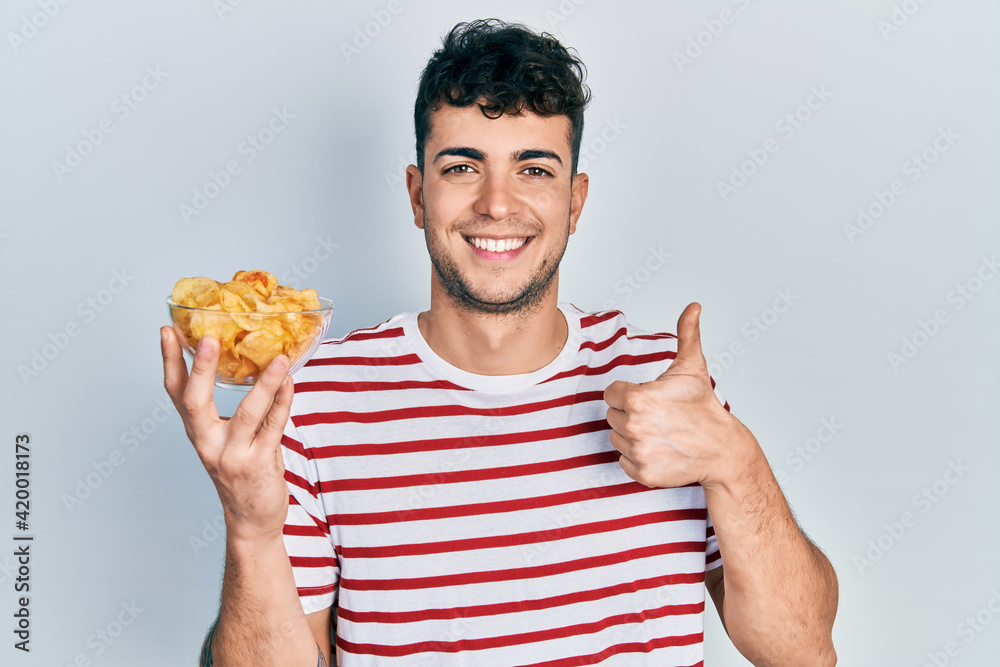 Young hispanic man holding potato chip smiling happy and positive, thumb up doing excellent and approval sign Young hispanic man holding potato chip smiling happy and positive, thumb up doing excellent and approval sign