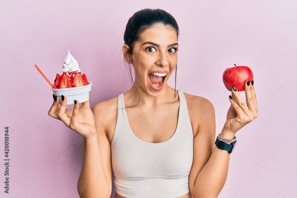 Young hispanic woman wearing sportswear holding apple and ice cream celebrating crazy and amazed for success with open eyes screaming excited. Young hispanic woman wearing sportswear holding apple and ice cream celebrating crazy and amazed for success with open eyes screaming excited.