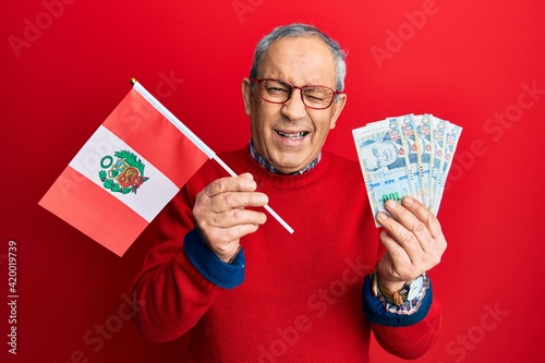 Handsome senior man with grey hair holding peru flag and peruvian sol banknotes winking looking at the camera with sexy expression, cheerful and happy face.