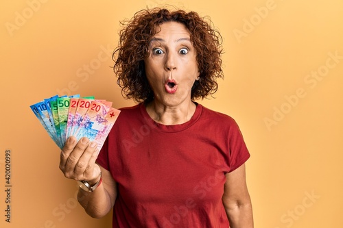 Beautiful middle age mature woman holding swiss franc banknotes scared and amazed with open mouth for surprise, disbelief face