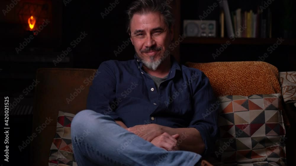 Portrait of older man at home in dark room sitting on couch. Happy smile, grey hair, bearded, . mature age, middle age, mid adult man in 50s. Portrait of older man at home in dark room sitting on couch. Happy smile, grey hair, bearded, . mature age, middle age, mid adult man in 50s.