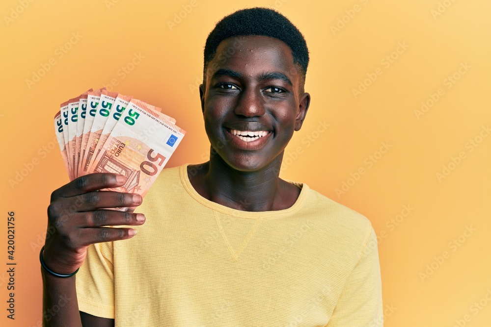 Young african american man holding 50 euro banknotes looking positive and happy standing and smiling with a confident smile showing teeth Young african american man holding 50 euro banknotes looking positive and happy standing and smiling with a confident smile showing teeth
