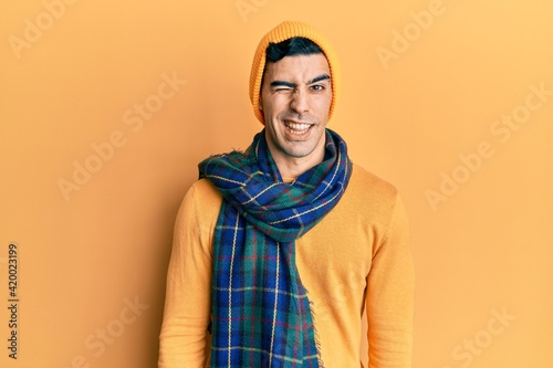 Handsome hispanic man wearing wool hat and winter scarf winking looking at the camera with sexy expression, cheerful and happy face. © Krakenimages.com